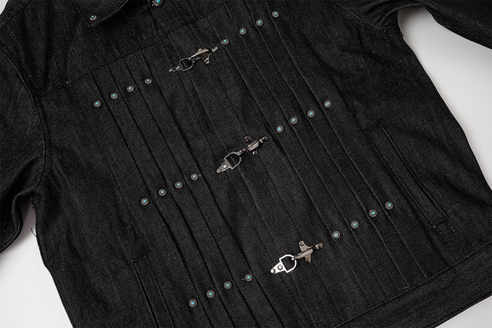 Close-up of the black denim jacket front showing vertical pleats, three metal clasp closures, and turquoise studs