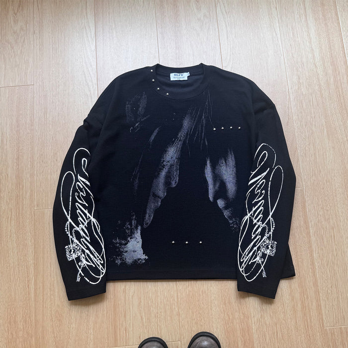 Front view of a black long-sleeve tee laid flat, featuring gray graphic artwork, white sleeve script, and studs