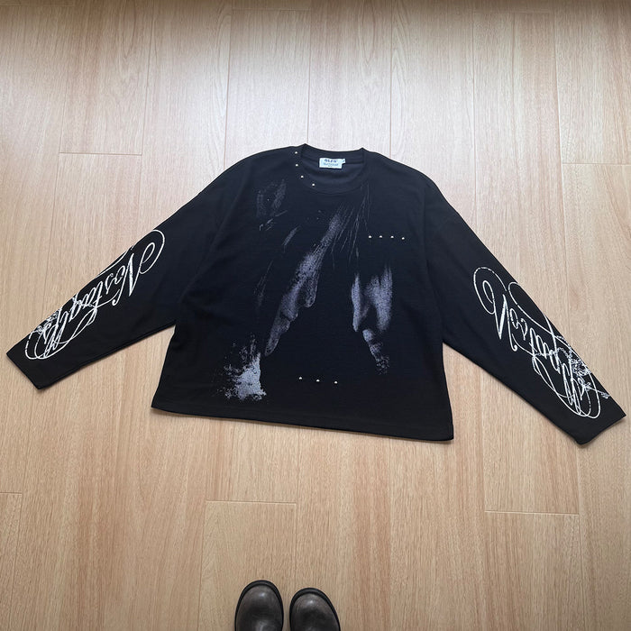 Black long-sleeve graphic tee laid flat with gray front artwork, white cursive sleeve prints, and metallic studs