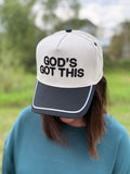 A person outdoors wearing a baseball cap with the text 'GOD'S GOT THIS' in black on an off-white crown and a black brim.