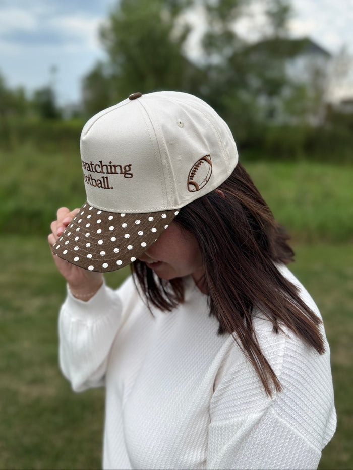 A woman outdoors wearing a cream baseball cap with brown polka-dot visor trim and brown embroidered text reading 'he's watching football.' on the front, paired with a white top.