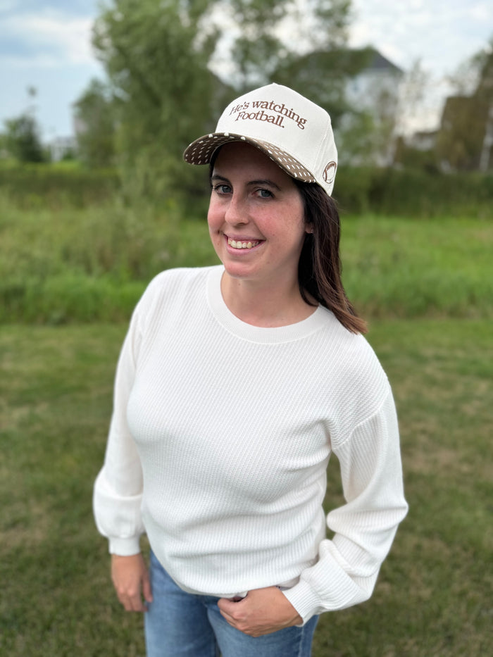 A woman standing outdoors in a grassy field, wearing a cream baseball cap with brown text that reads 'He's watching football'.