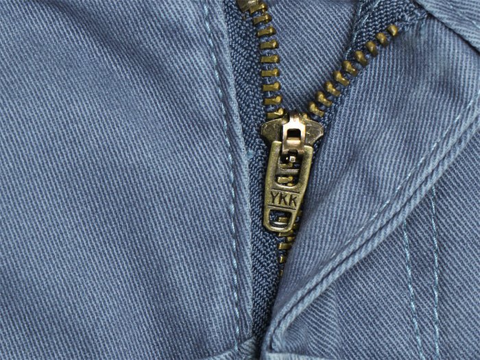 Macro view of the blue pants fly showing a brass-toned zipper, woven fabric texture, and reinforced stitching