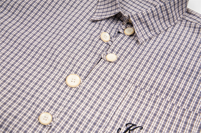 Close-up of the light beige plaid shirt front showing cream buttons, collar, and partial embroidered chest detail