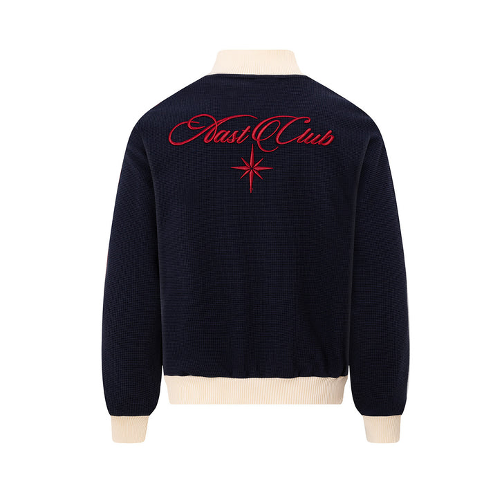 Back view of a navy waffle-knit track jacket with cream ribbed collar, hem and cuffs, plus red embroidered logo