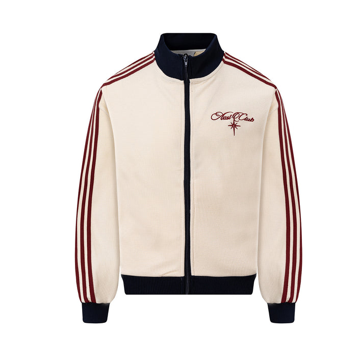 Front view of a cream waffle-knit track jacket with navy collar, zipper, hem and cuffs, red stripes and chest embroidery