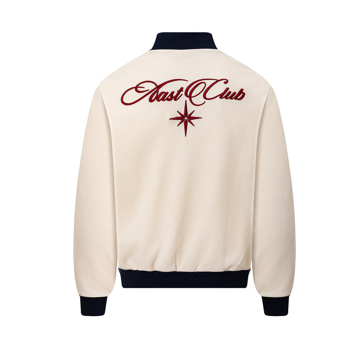 Back view of a cream waffle-knit track jacket with navy ribbed collar and hem, featuring large red embroidered lettering