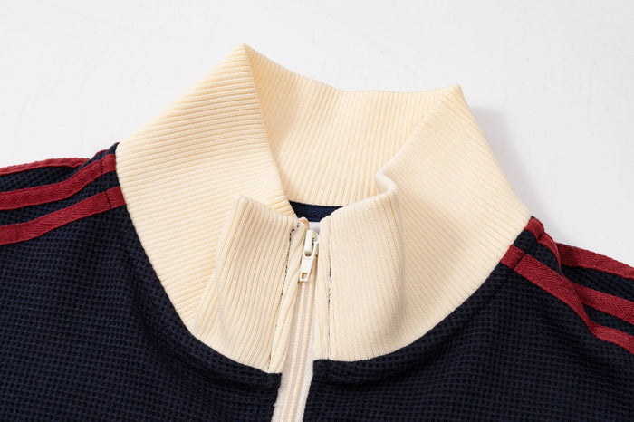 Close-up of a navy track jacket showing its cream ribbed collar, front zipper and red shoulder stripes