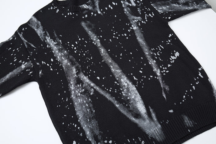 Black knit sweater laid flat, showing the front paint-splatter pattern and ribbed hem