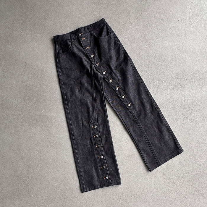 Flat lay of raw black denim jeans showing the front, straight legs, waistband, pockets, and side snaps