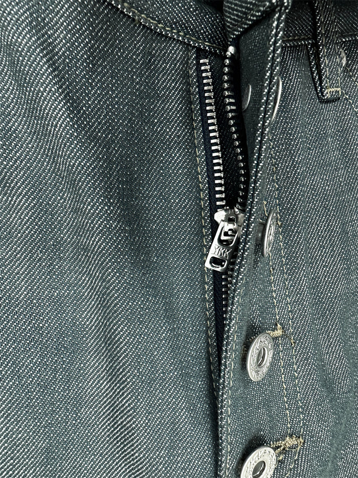 Close-up of lake green jeans showing the metal zipper, button fly edge, rivets, and diagonal denim weave